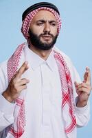 Arab man dressed in cultural thobe and checkered headscarf frowning eyebrows and crossing fingers while making wish. Superstitious muslim person pleading for luck with gesture photo