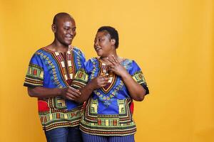 Smiling man and woman romantic pair chatting while standing in studio. Happy wife and husband wearing ethnic african american clothes talking and posing on orange background photo