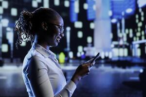 BIPOC woman walking around city at night, scrolling on online shopping platform using mobile phone. Citizen using smartphone while strolling outside on empty streets illuminated by lamps photo