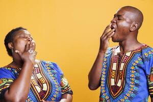 Tired black man and woman pair yawning and covering mouth with hand. Exhausted african american couple showing lack of sleep gesture and feeling fatigue in morning in studio photo