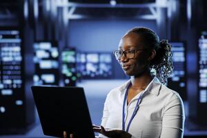 Relaxed african american admin uses laptop to prevent system overload during peak traffic periods. Woman between server clusters ensuring enough network bandwidth for smooth operations photo
