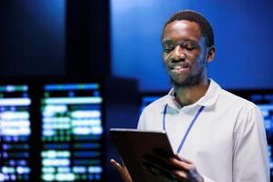 African american serviceman using laptop to review energy consumption across server cabinets electronics. Happy supervisor ensuring data center equipment temperature sensors are performing flawlessly photo