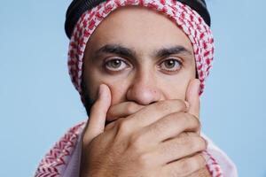 Muslim man covering mouth with palms while keeping silence and looking at camera closeup. Arab person holding hands on lips while showing speak no evil three wise monkeys concept photo