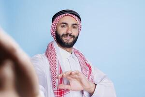 Smiling arab man wearing traditional attire holding mobile phone in arm and looking at front camera fpv. Muslim person with cheerful expression talking in videocall pov perspective photo