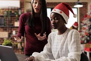 African american woman employee in santa hat working with task on laptop and getting help from colleague. Coworkers brainstorming and managing project report in office with christmas setting photo