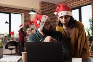 Colleagues discussing and solving task on laptop while coworking in office during new year winter season. Caucasian man and woman coworkers in santa hats checking report on computer photo