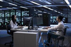 Person with paraplegia and colleague working in inclusive server hub workspace, typing on PC. Man in wheelchair and coworker doing tasks in data center housing advanced data storage infrastructure photo