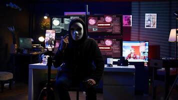 Hacker concealing identity filming ransom in hidden underground base, threatening to release stolen data publicly if payout demands are not met, using phone to record blackmail footage, camera B photo