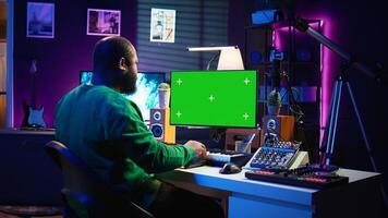 Songwriter operates on synthesizer piano keys next to isolated display, composing new music with instruments and stereo tools in home studio. Artist producer works with green screen. Camera B. photo