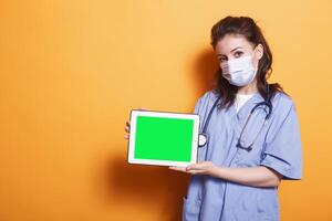 Nurse practitioner with face mask horizontally holding tablet with green screen. Female healthcare specialist grasping digital device displaying isolated copyspace mockup template. photo