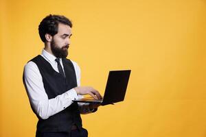 Restaurant worker checking website on laptop, browses online in studio to create new price list on restaurant menu. Young adult uses pc to navigate through social media app on camera. photo