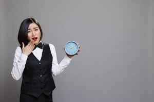 Tired asian woman wearing waitress uniform yawning and covering mouth while holding alarm clock portrait. Sleepy receptionist running late for work, showing time and looking at camera photo