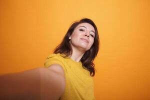 POV of caucasian female individual taking a selfie in studio. Cheerful woman smiling and standing over isolated background, making a personal vlog. Casual person taking picture with smartphone. photo