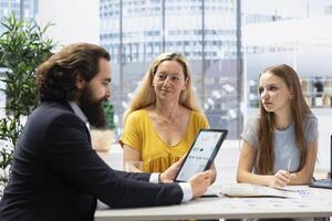 Financial advisor creating reports and statistic charts on investment performance on tablet, showing them to clients. Agent using device to develop portfolio diagram graphs for customers photo