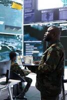 Team of military specialists overseeing surveillance activity by using a cctv footage and radar, pointing at a big screen and world map. High tech control center base of operations. photo