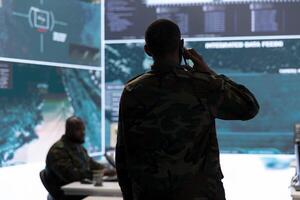 Military man answers phone call in a mission control center, working with advanced technology and satellite imagery to track potential threats and monitor real time data. Intelligence activities. photo