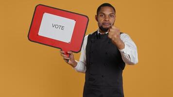 Waiter does an advertisement for politics and showing a speech bubble with the message to vote, standing against yellow background. Restaurant staff holds a cardboard sign. Camera B. photo
