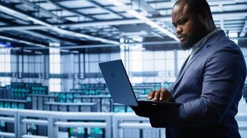 Software developer in server hub checking recovery plan on laptop, monitoring hardware energy consumption. African american worker uses notebook in data center, ensuring rigs are functioning, camera B photo