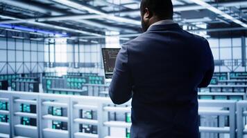 African american admin in data center running error checking utilities on tablet to fix hardware bugs. IT expert in server hub workspace looking for issues affecting mainframes performance, camera B photo
