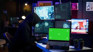 Hacker in hidden place using green screen laptop to deploy malware on unsecured devices, stealing sensitive data from unaware users online and selling it on black market, camera B photo