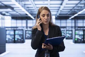 Manager maintaining accurate documentation of data center equipment, chatting on mobile phone with employee. Woman talks about server farm configurations and procedures during telephone call photo