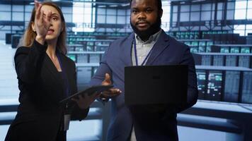 Teamworking colleagues doing checkup, inputting data on laptop of needed server hub fixes. Coworkers working together in data center housing advanced storage infrastructure, camera A photo