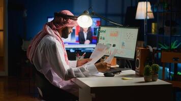 Arabic teleworker checking analytics statistical data set while remotely working in apartment office. Company executive optimizing key performance indicators with tv as background noise photo