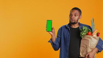 Young guy with a bag of fresh groceries showing greenscreen on his smartphone, presenting an isolated display on phone. Person advertising locally grown produce, zero waste. Camera B. photo