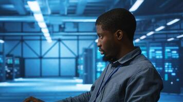 African american IT expert in server farm ensuring valuable data remains shielded from threats. Man safeguarding data center hardware against unauthorized access and vulnerabilities, securing system photo