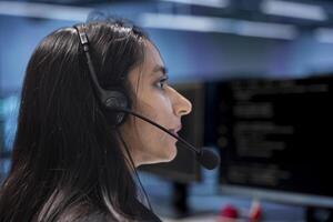Admin in data center facility managing call center tasks, speaking into headset microphone while monitoring multiple PC screens. Indian woman talking with customers through headphones mic photo