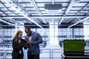 Software developers using green screen laptop in data center, making sure rigs are functioning optimally. Experts in server room using chroma key laptop to monitor energy consumption photo