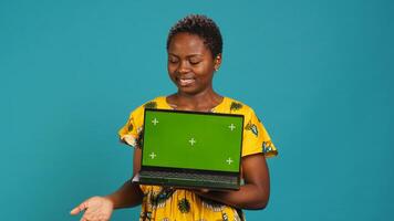 Confident smiling woman presenting a chroma key display on laptop, standing against blue background. Young adult holding a wireless pc and showing a green screen mockup. Camera A. photo