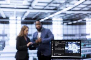 Focus on laptop in data center used by team of specialists in blurry background updating AI systems. Close up of device used by colleagues upgrading artificial intelligence simulation model photo