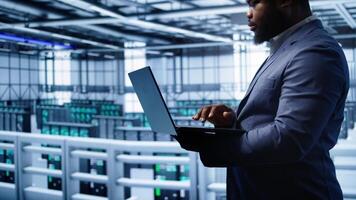 Software developer in server hub checking recovery plan on laptop, monitoring hardware energy consumption. African american worker uses notebook in data center, ensuring rigs are functioning, camera B photo