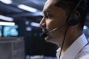 Data center agent assisting engineering teams with hardware installations, issuing commands though headset mic. Close up of IT specialist in server farm communicating with engineers in remote call photo