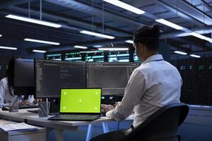 Engineer in server hub using isolated screen laptop to repair equipment storing datasets. Professional using chroma key notebook to do debugging on rigs doing computational operations photo