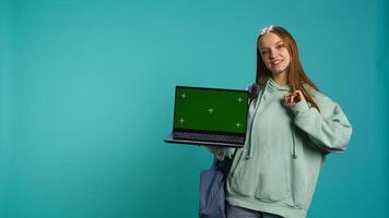 Happy girl presenting laptop with green screen display, isolated over studio backdrop. Radiant teenager creating promoting advertisement with mockup notebook device, camera B photo