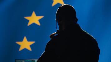 European Union military dispatcher looking at surveillance broadcast, relaying informations for troops using headset. Telecommunications operator over EU flag background, coordinating troops, camera A photo