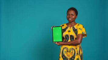 Feminine person in native clothing holds a tablet with mockup screen, creating an advertisement against blue background in studio. Young woman smiling and presenting copy space. Camera B. photo