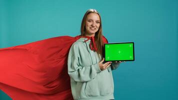Joyous girl portraying superhero with red cape showcasing green screen tablet, studio backdrop. Joyful teenager posing as hero in costume on advertising chroma key digital device, camera B photo