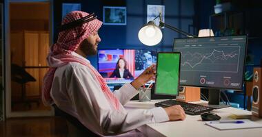 Muslim remote worker at home holds chroma key tablet while looking at business annual report graphs on computer. Arabic freelancer with mockup digital device in hand performing company tasks photo