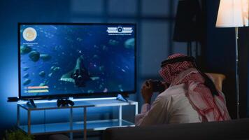 Arabic gamer playing classic arcade action videogame, flying through space debris. Muslim man enjoying leisure time at home using high tech gaming console to solve missions in singleplayer game photo