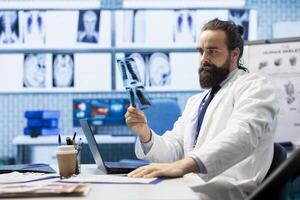 General practitioner inspecting x ray scans and test results in medical clinic, working with hospital records on laptop to ensure the best treatment plan for disease prevention. Radiology expert. photo