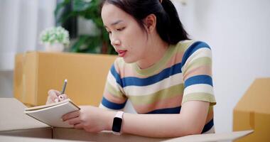 Handheld shot, Young Asian woman is taking notes while checking belongings in cardboard boxes in preparation for moving to a new house. Moving house and lifestyle concept. video