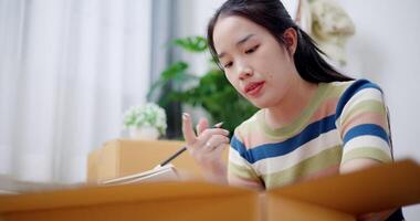 Handheld shot, Young Asian woman is taking notes while checking belongings in cardboard boxes in preparation for moving to a new house. Moving house and lifestyle concept. video