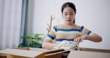 Handheld selective focus shot, Young Asian woman is sitting on the floor folding clothes into a cardboard box to prepare for a move. Moving house and lifestyle concept. video