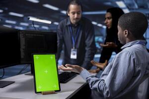 Focus on green screen tablet used by team in blurry background reconfiguring server farm to optimize it for data processing. Close up of mockup device used by workers overseeing data center equipment photo