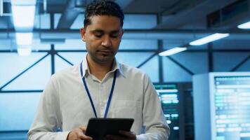 Programmer walking in high tech data center used for managing infrastructure energy consumption, working on tablet. Indian man using device in workspace used for automation in server farm, camera A photo