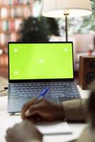 Entrepreneur using a notebook to write notes next to green screen display on laptop, working from home to help develop the startup. Businessman analyzes accounting data at desk. photo