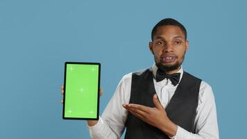 Bellman hotelier showing a tablet with green screen display in studio, presenting mockup chroma key screen against blue background. Young hotel concierge works at resort. Camera A. photo